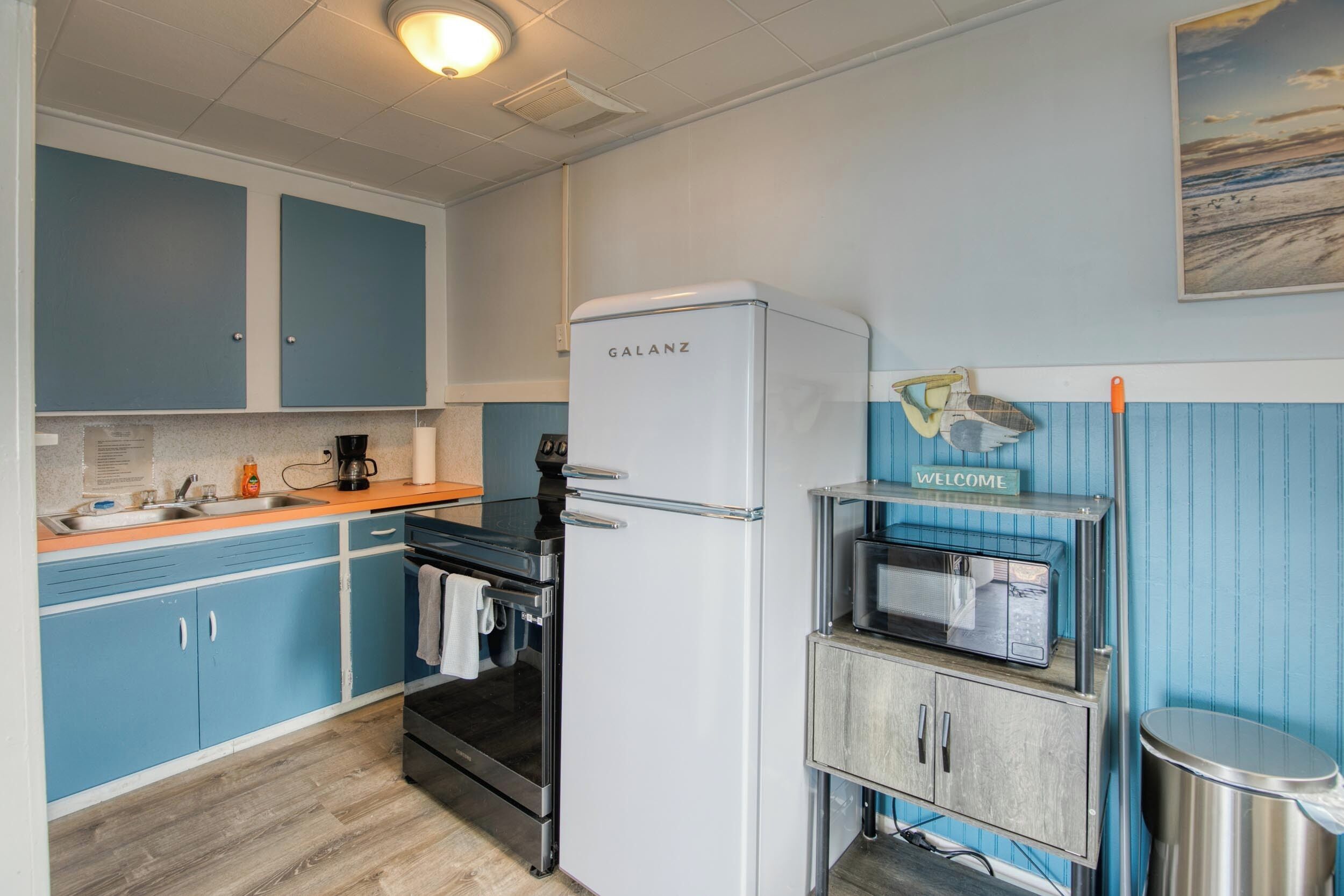 A compact kitchen with blue cabinets, a white refrigerator, a stove, and a microwave on a wooden shelf.