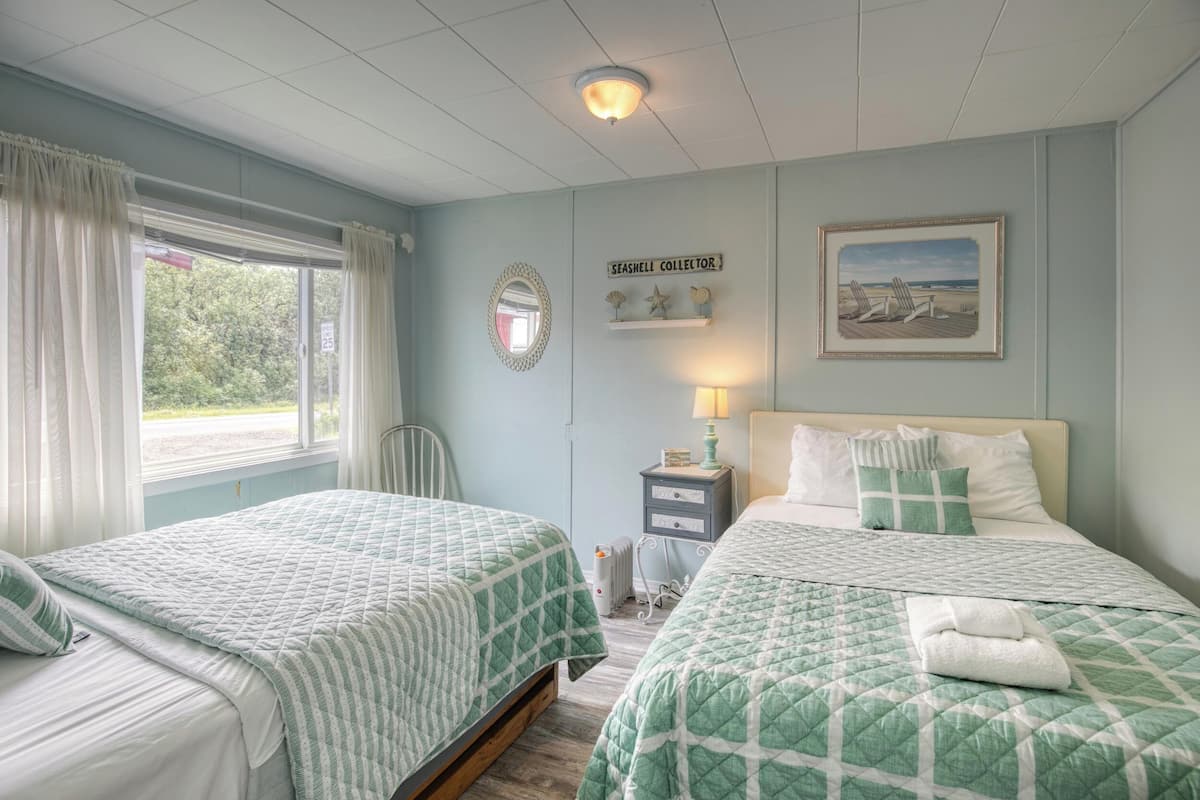 A bedroom with two beds, a window with curtains, a bedside table with a lamp, a framed picture on the wall, and a sign above the bed.