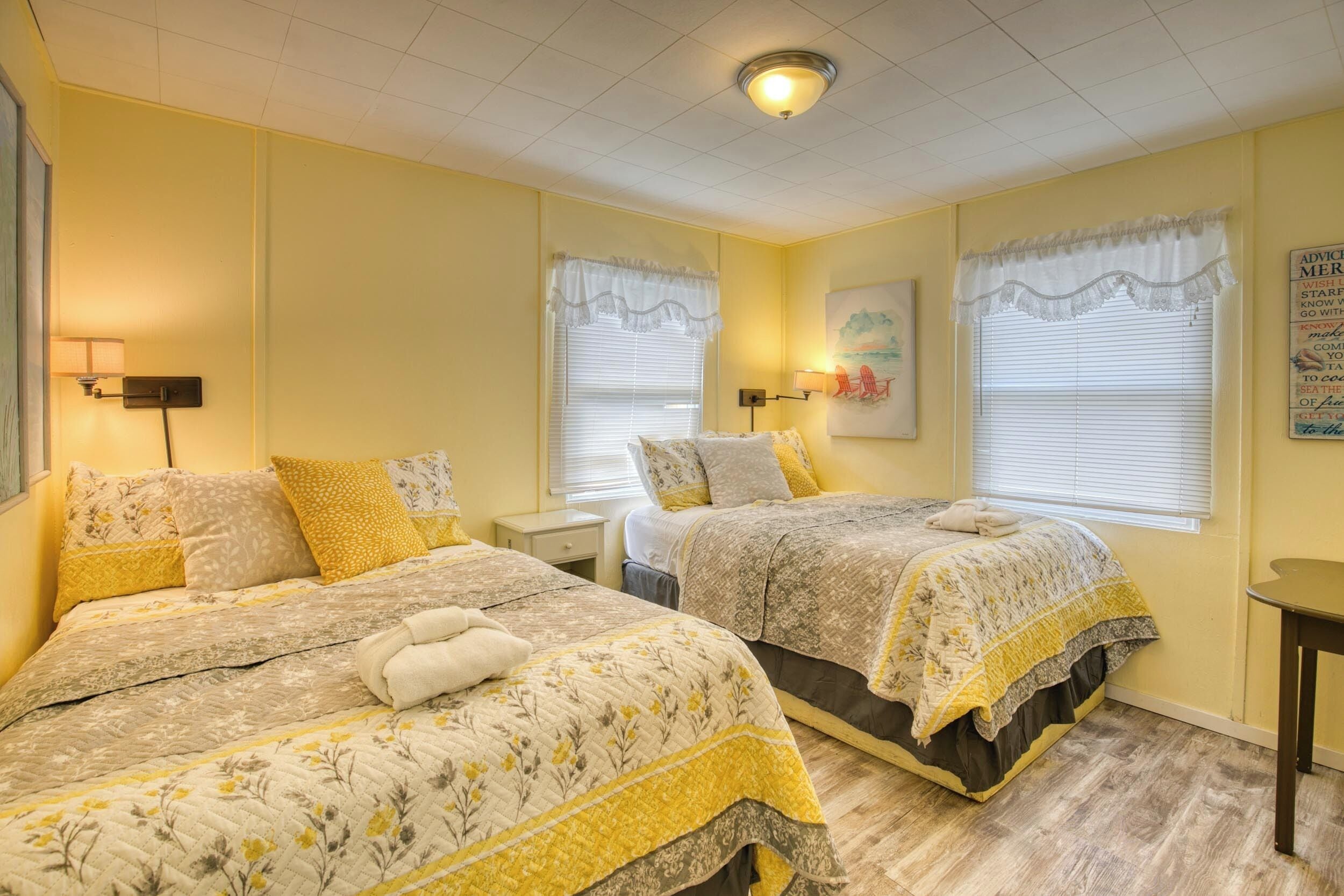 Habitación de hotel con dos camas, cada una con ropa de cama y almohadas de estampado floral, una mesita de noche con lámpara y un ventanal con cortinilla blanca.