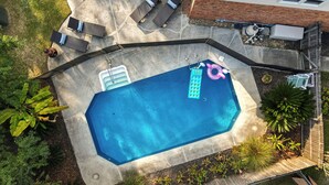 Oversigtsbillede af en baghave med swimmingpool, terrasse med liggestole og et murstenshus.
