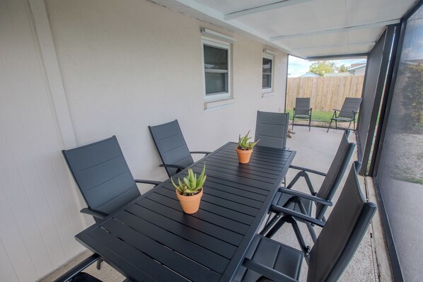 Un patio techado con una mesa y sillas de metal negro, dos plantas en macetas y una valla de madera al fondo.