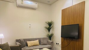 A living room with a grey sofa, a coffee table, a TV mounted on a wooden panel, and a patterned floor. - Lordy Serviced Apartments (Riyadh)