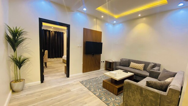 A modern living room with a grey sofa, a coffee table, and a TV. There is a wooden floor and a potted plant. - Lordy Serviced Apartments (Riyadh)