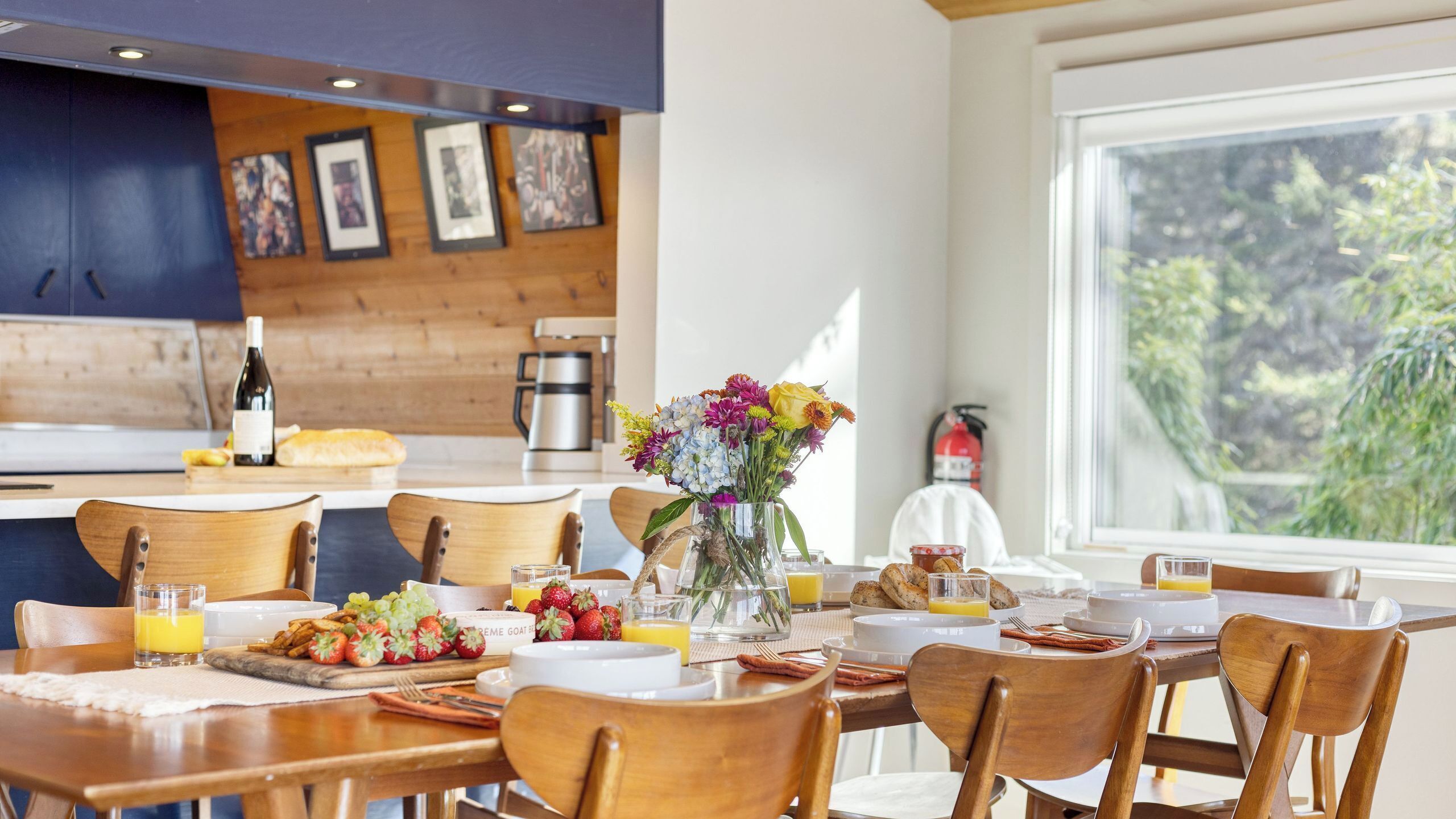 A dining area with wooden chairs, a table set with breakfast, a vase with flowers, and a view of greenery outside.
