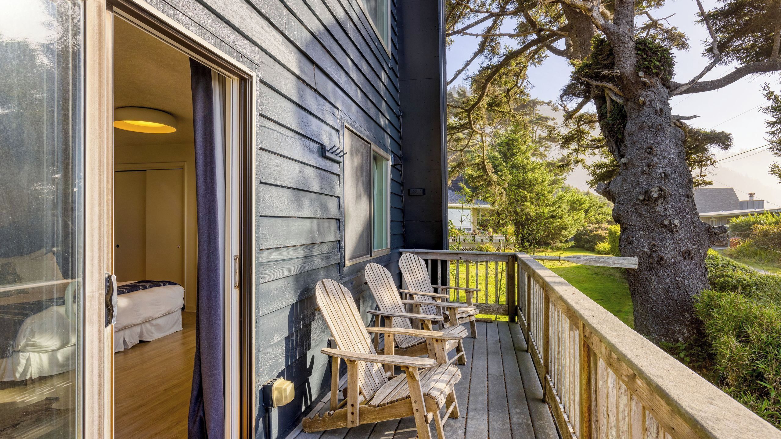 Un pontile in legno con poltrone Adirondack, una porta vetrata che dà su una stanza con un letto e vista su un albero e il paesaggio.