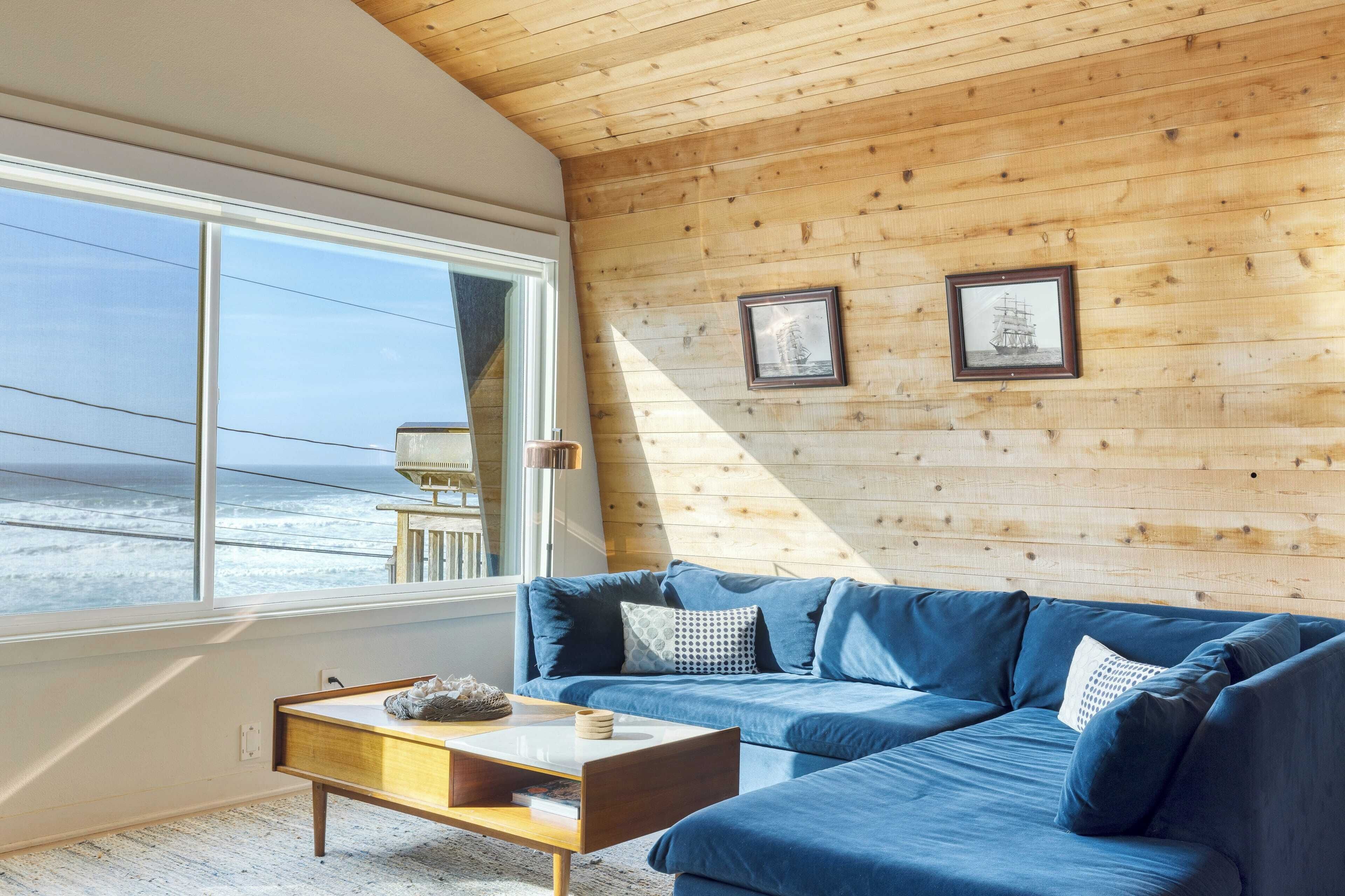 A cozy room with a blue sofa, wooden walls, and a large window overlooking the ocean.