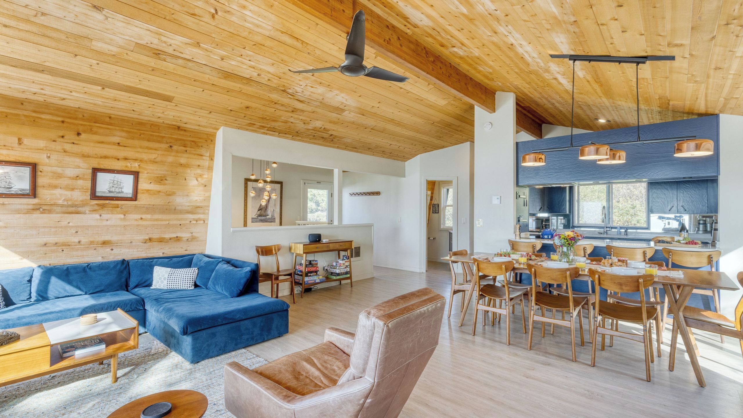 A spacious living area with a blue sofa, wooden dining table, and chairs, a kitchen with dark cabinets, and a ceiling fan.