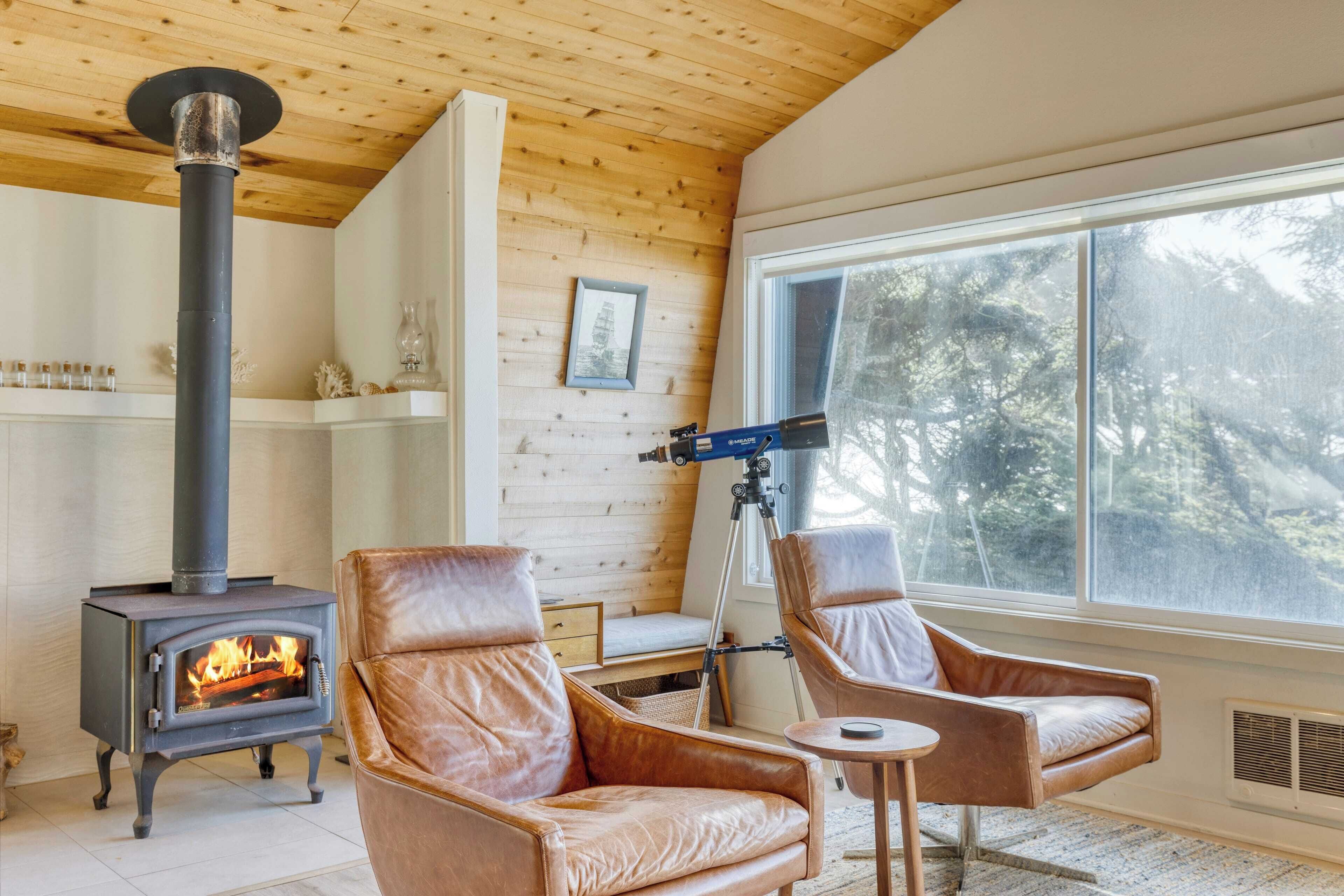 A cozy room with a wood-burning stove, leather chairs, a telescope, and a large window overlooking trees.