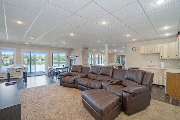 A spacious living area with a large sectional sofa, a fireplace, and a view of a lake through large windows. - Stunning Lake House - 200` of Lakefront with Kayaks & Game Room (Kaleva)