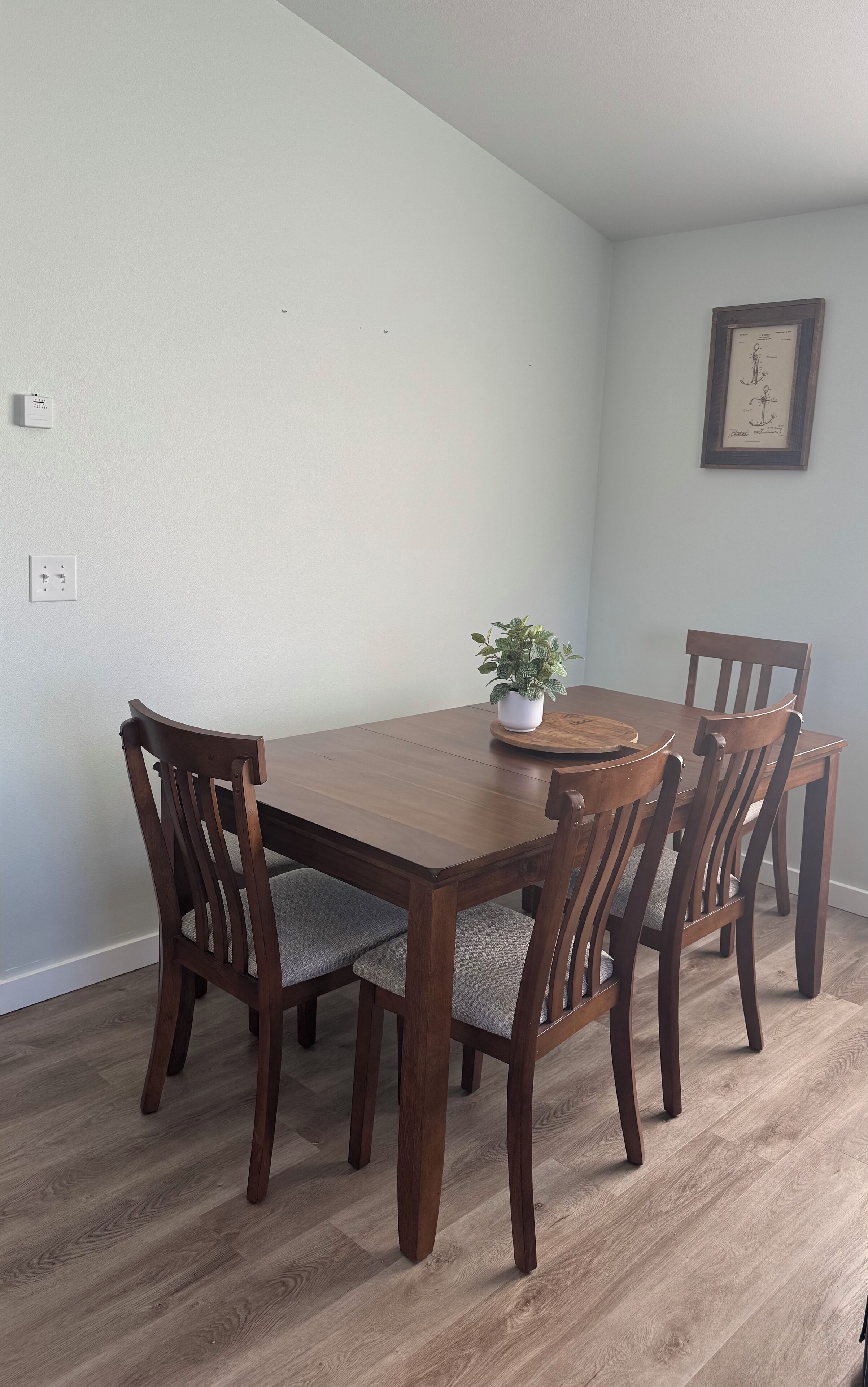 A dining room with a wooden table and chairs, a potted plant, and a framed picture on the wall.