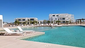 Ein moderner Poolbereich mit Liegestühlen und einem klaren, blauen Pool, umgeben von weißen Gebäuden und Palmen.
