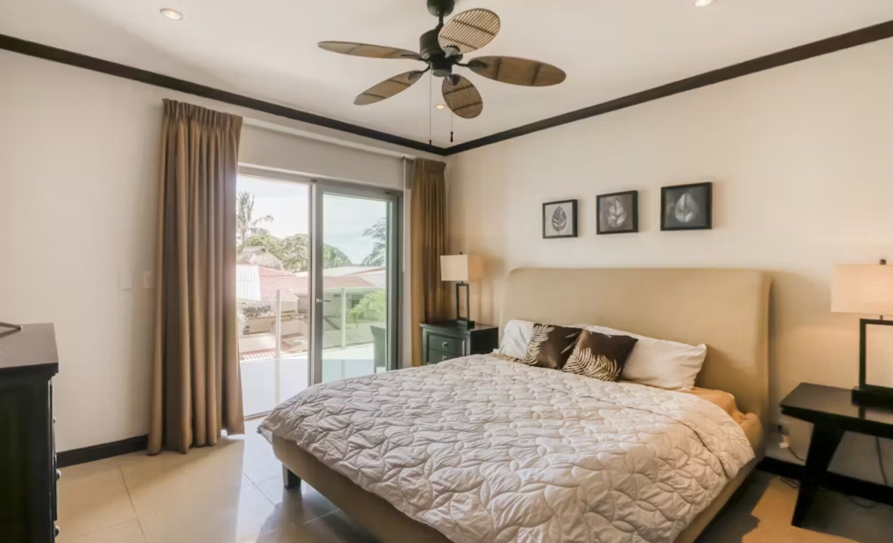 Une chambre d’hôtel avec un grand lit, un ventilateur de plafond, une vue sur l’extérieur grâce à une porte-fenêtre coulissante, et un petit bureau avec une lampe.