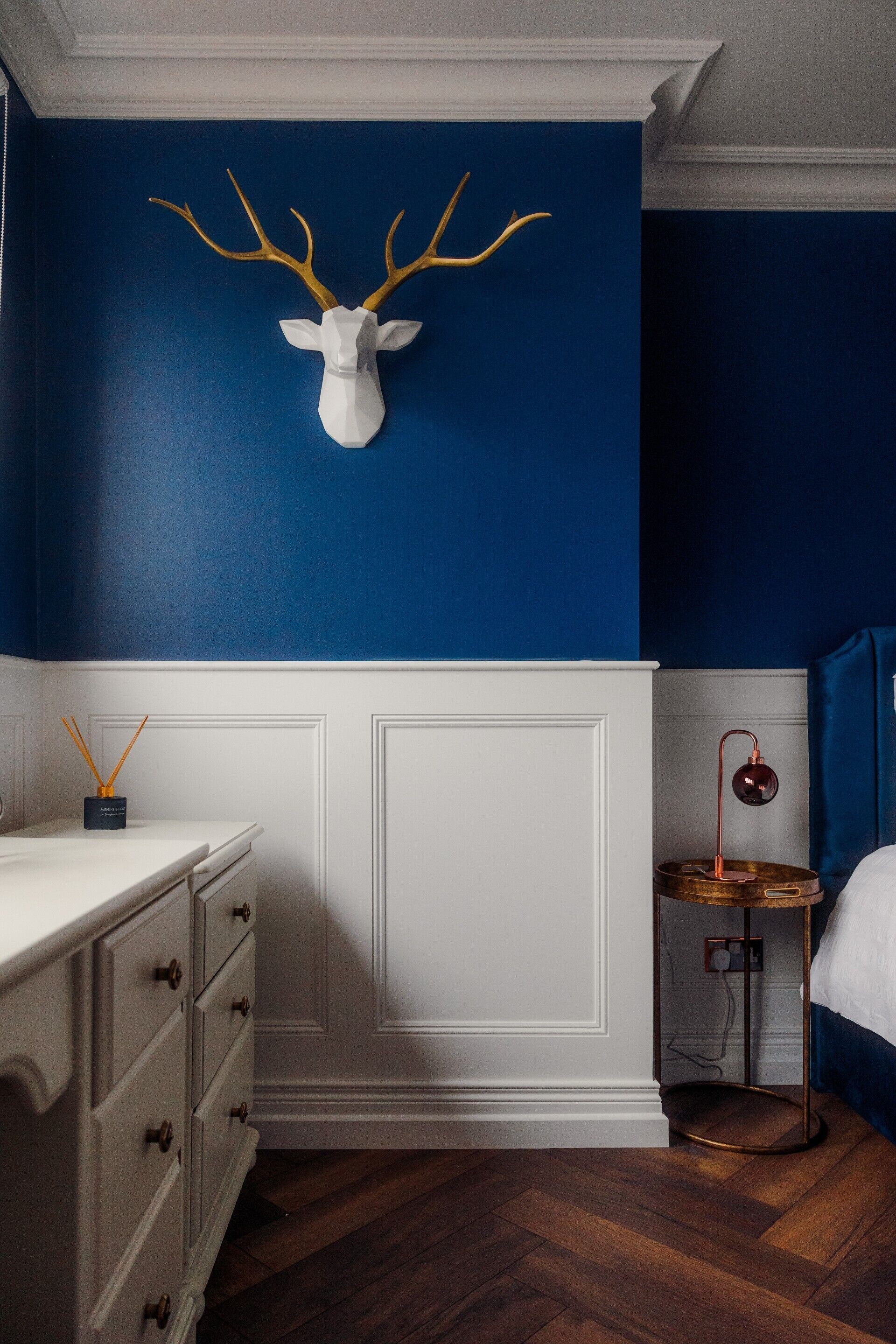 A bedroom with a blue wall, a white dresser, a round table with a lamp, and a mounted deer head.