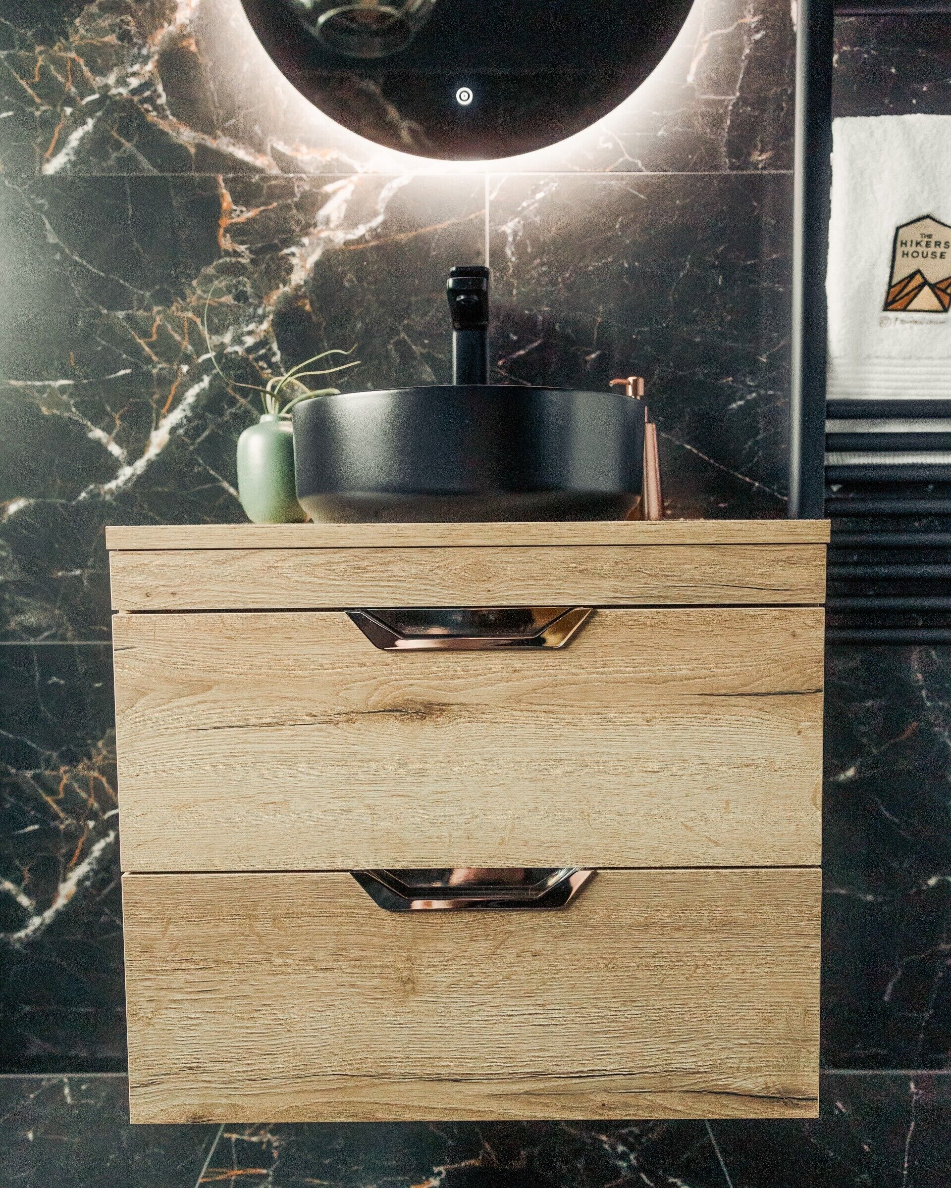 Een houten wastafelmeubel met een zwarte wasbak, een groene vaas en een logo op de muur.
