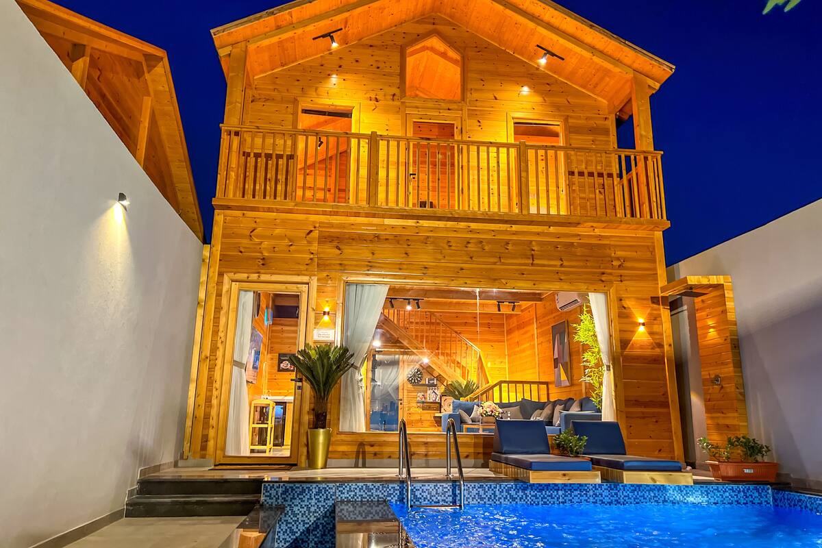 Una cabaña de madera de dos pisos con un balcón, una piscina y una escalera que lleva al piso superior.