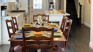 On aperçoit une salle à manger avec une table en bois dressée pour six personnes, entourée de chaises en bois. Au fond, on distingue une cuisine avec des armoires blanches et un réfrigérateur en acier inoxydable.