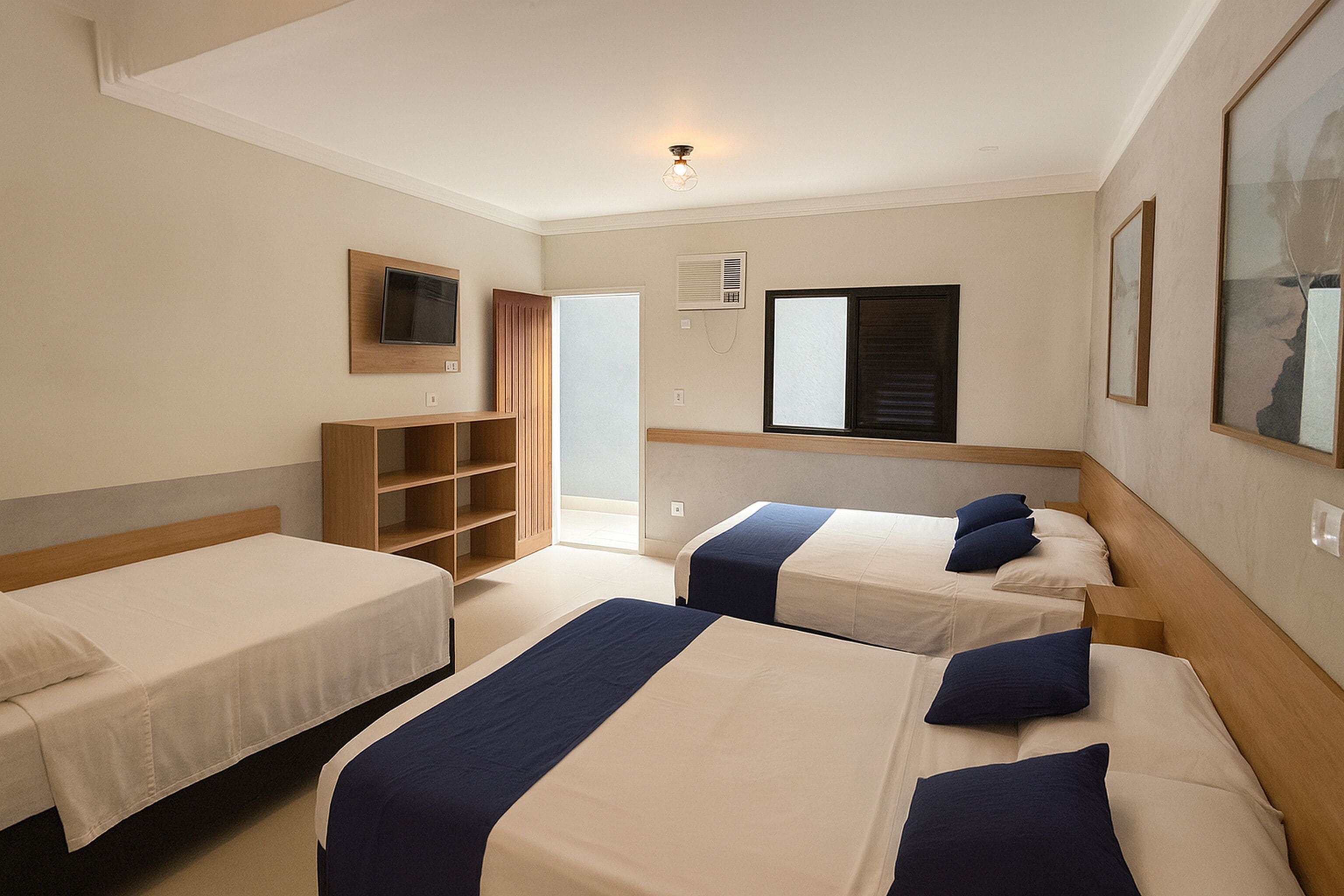 Habitació d'hotel amb tres llits, una televisió penjada a la paret, una finestra amb persianes i una porta que dona a un balcó.