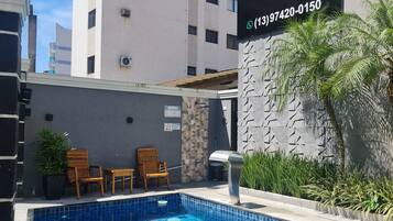 A swimming pool with blue tiles, surrounded by a lounge area with wooden chairs and a potted plant. A sign for Marmeu Apartment Hotel is visible in the background.