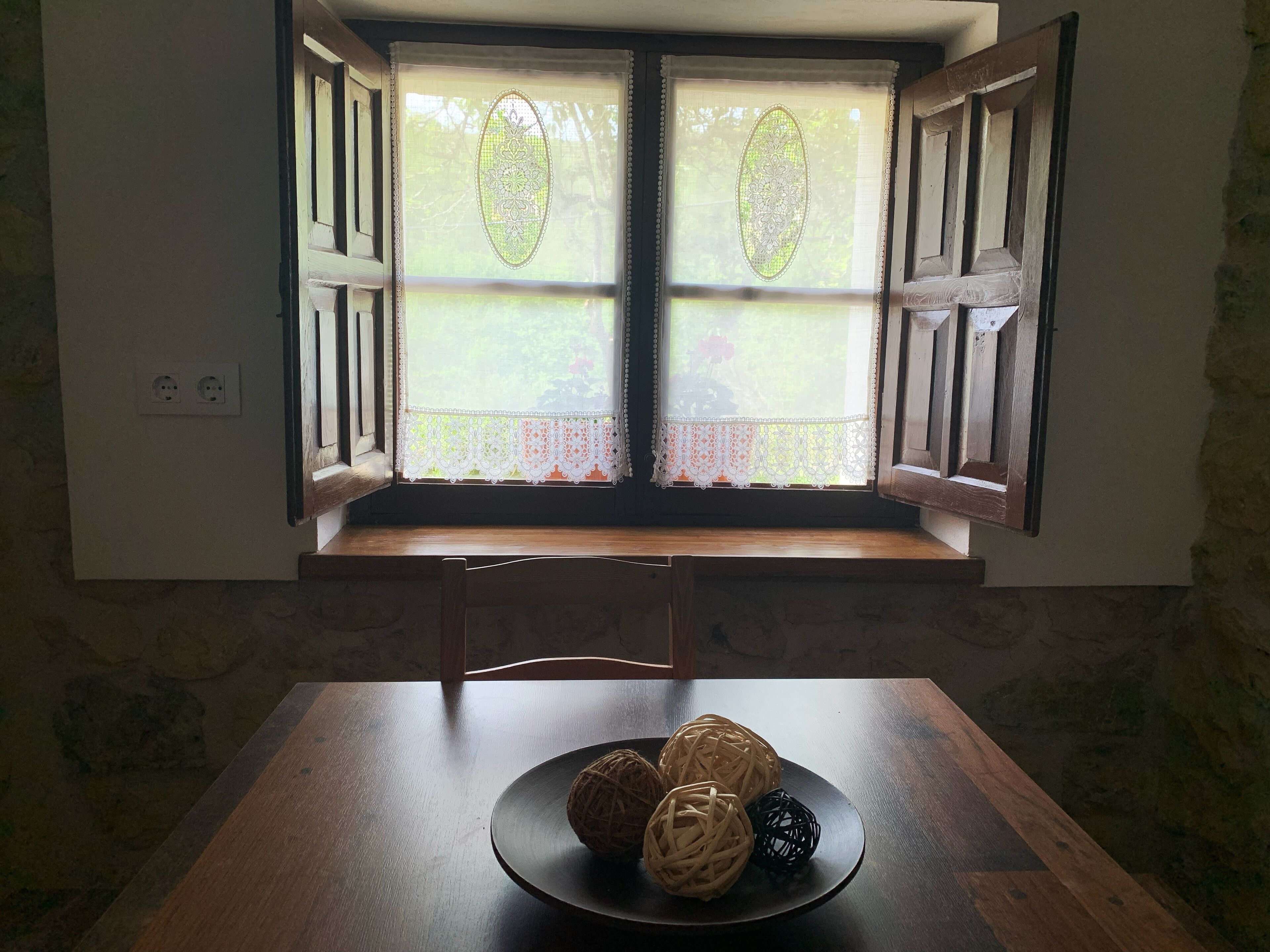 Una habitación con una mesa de madera, una ventana con contraventanas abiertas y vistas a la vegetación.