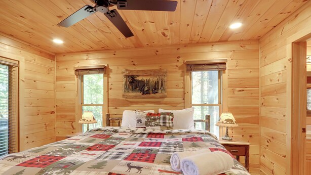 Quarto de cabana de madeira com cama, duas mesas de cabeceira, ventilador de teto e janela com vista para árvores.