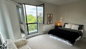 A bedroom with a large window, a bed with a white blanket, a bedside table with a lamp, and a radiator. - The Charm London  (Londres)
