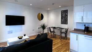 A modern living room with a black sofa, a dining area with a glass table, and a kitchen with white cabinets.