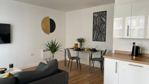 A modern living room with a sofa, a dining area with a table and chairs, a kitchen with cabinets and a coffee maker, and a TV mounted on the wall.