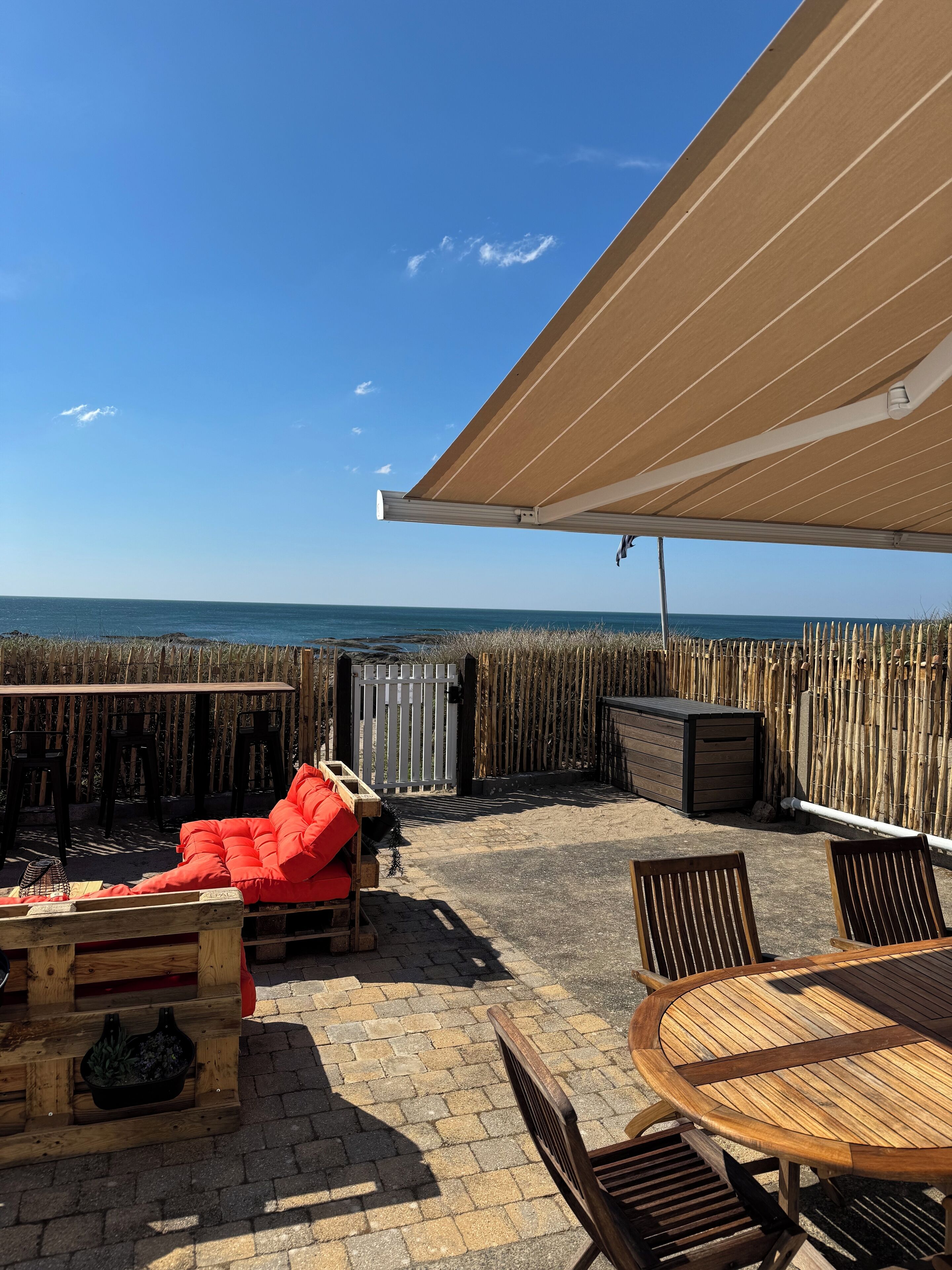 An outdoor patio with a wooden table, chairs, and a red sofa. A wooden fence and a wooden chest are also visible.