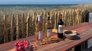 Une table en bois, sur laquelle sont disposées des bouteilles de vin, un bol de noix et un panier de fruits, donnant sur une plage bordée d’une clôture en bois.