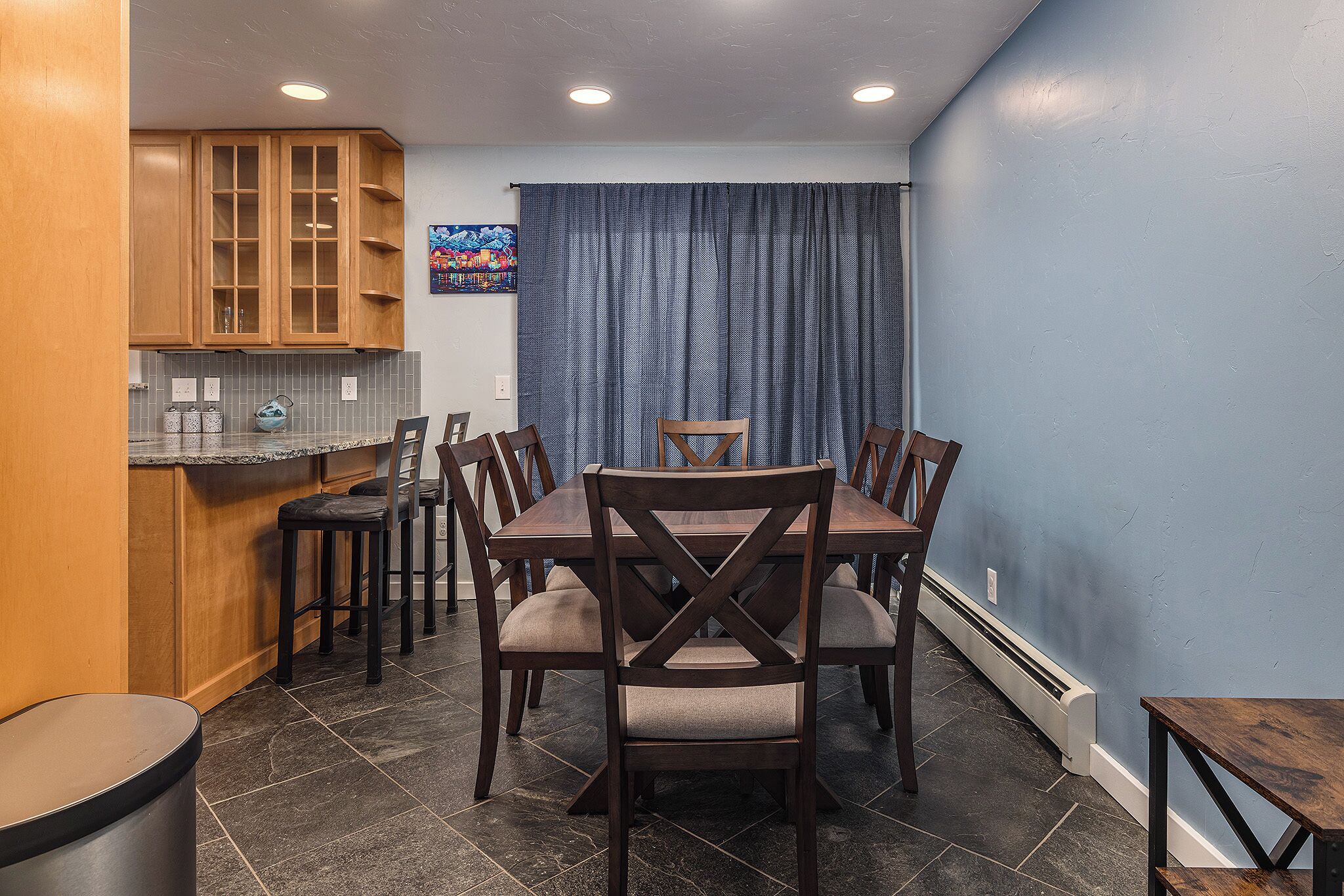 A dining area with a wooden table and chairs, a kitchen with wooden cabinets, and a blue wall.