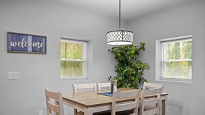 Un espace repas avec une table et des chaises en bois, un luminaire suspendu, un panneau d’accueil et une plante en pot.