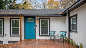 Une maison avec une porte bleue, deux fenêtres avec volets, un porche avec des chaises et une terrasse en bois.