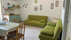 Un saló amb un sofà verd, una taula de dinar amb cadires, una zona de cuina i una taula petita amb objectes decoratius.
