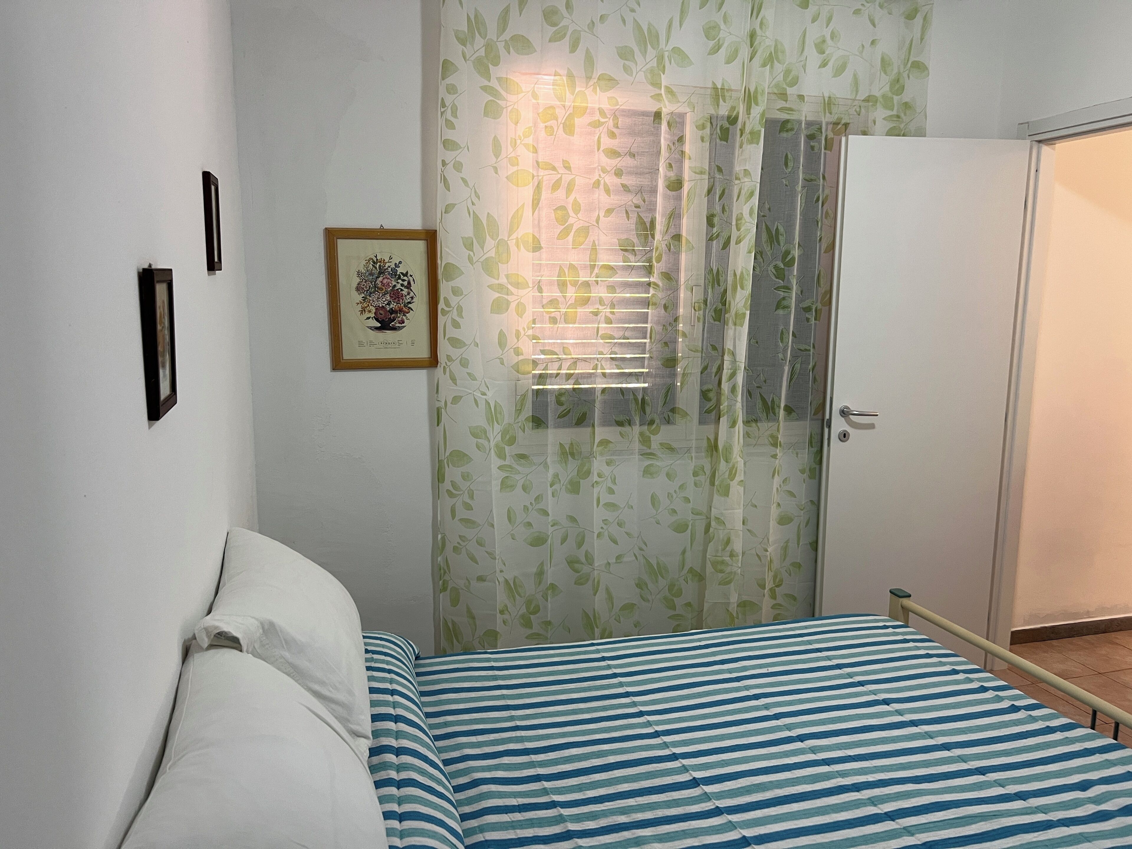 A bedroom with a bed, a wardrobe, a framed picture on the wall, and a window with a leaf-patterned curtain.