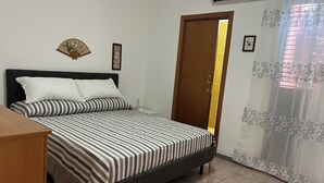 Un dormitori amb un llit, una petita taula de nit de fusta, un escriptori, una porta i un aparell d'aire condicionat.