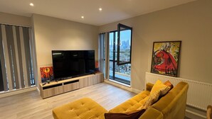 A modern living room with a large flat-screen TV, a yellow sofa, and a city view through large windows. - The Charm view London  (Londres)