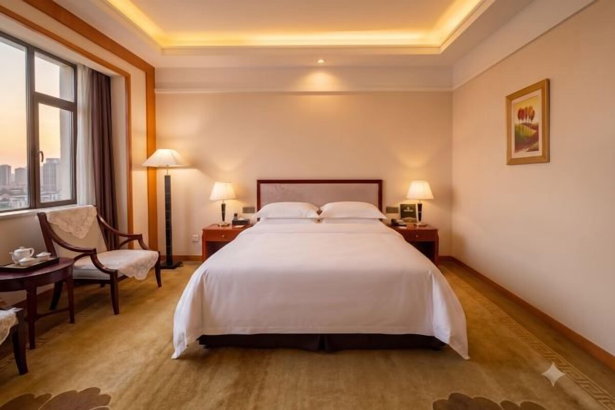 Une chambre d’hôtel avec un grand lit, des tables de chevet avec des lampes, une chaise, une fenêtre donnant sur un beau paysage et un tableau encadré au mur.