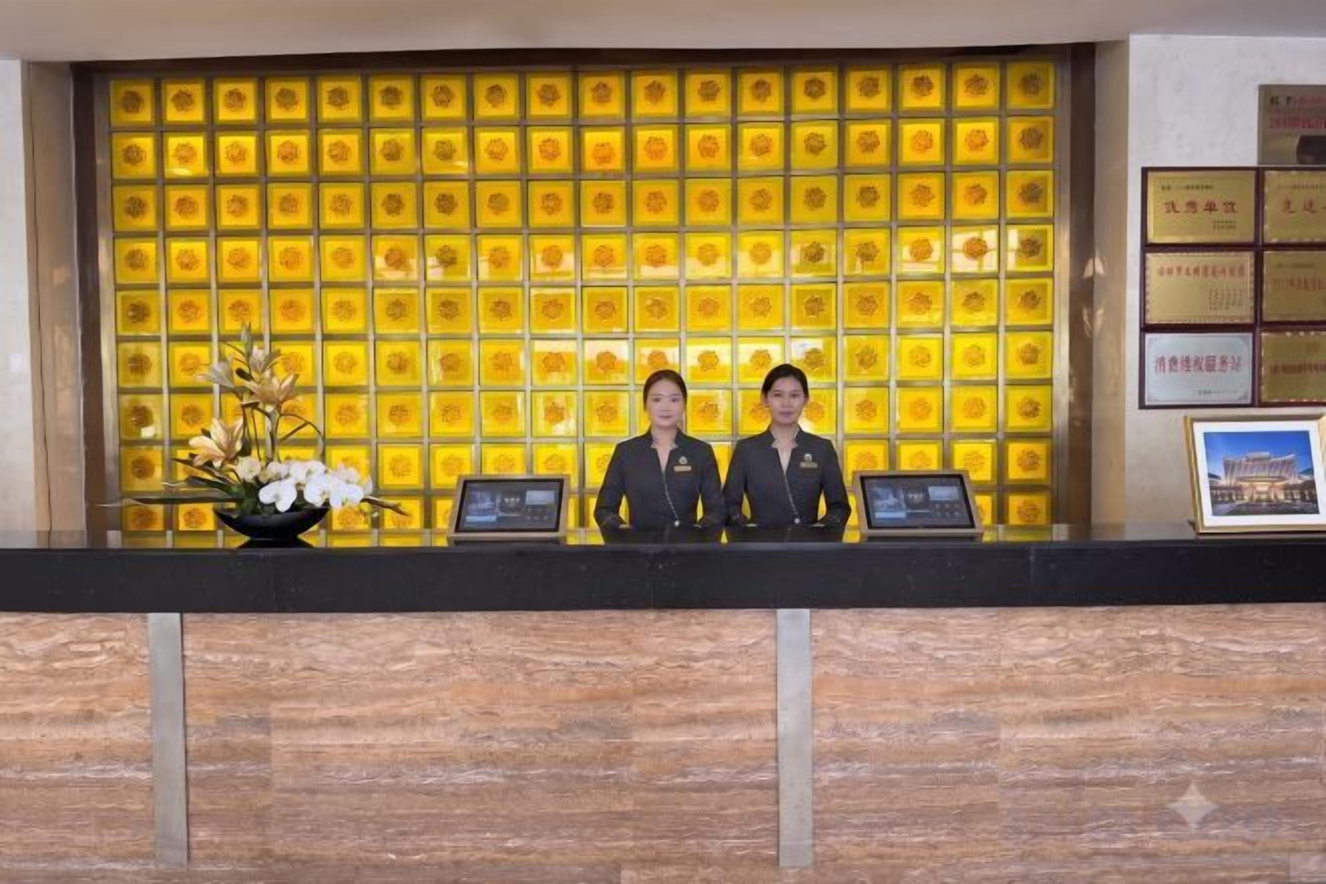 A reception desk with two staff members, a yellow patterned wall, and a framed picture on the wall.