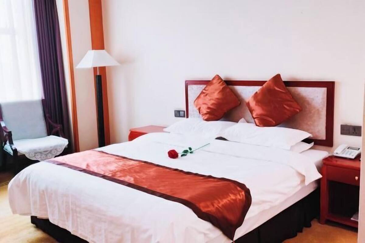 A neatly made bed with a white and brown bedspread, two red pillows, and a single red rose.