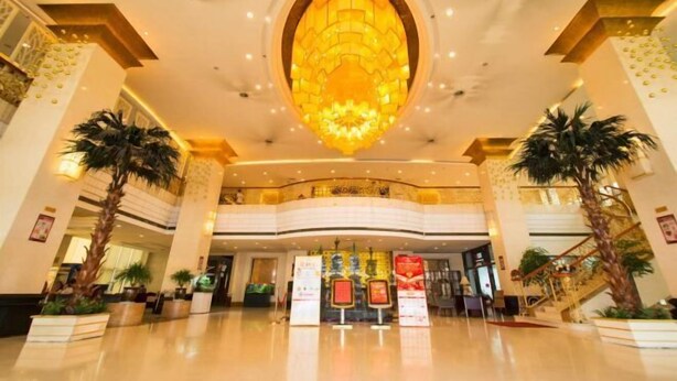 A spacious lobby with a large, ornate chandelier, palm trees, and a central reception area with informational displays. - Tianrui Zhongzhou International Hotel (Ruzhou)