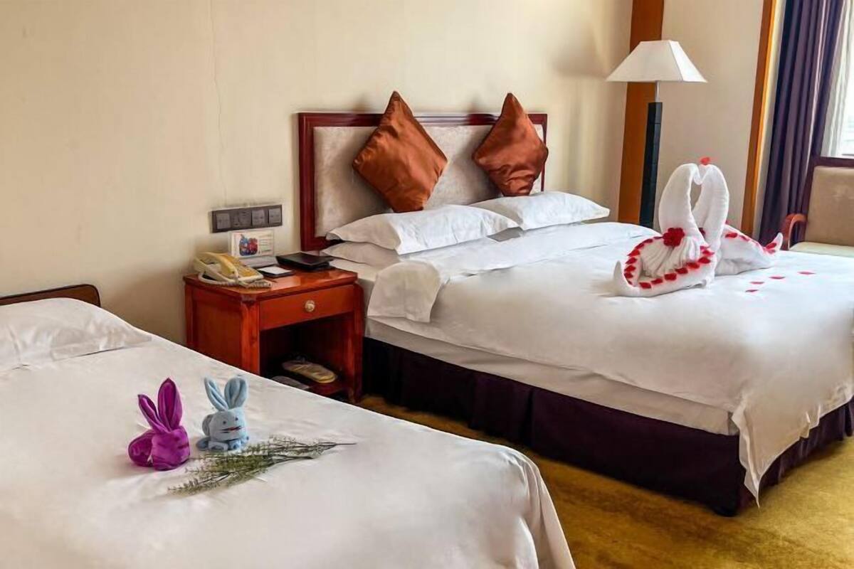 A hotel room with two beds, each with white linens and brown pillows. There are decorative items on the beds, including a towel sculpture and a small bunny figure. A nightstand with a phone and a lamp is visible.