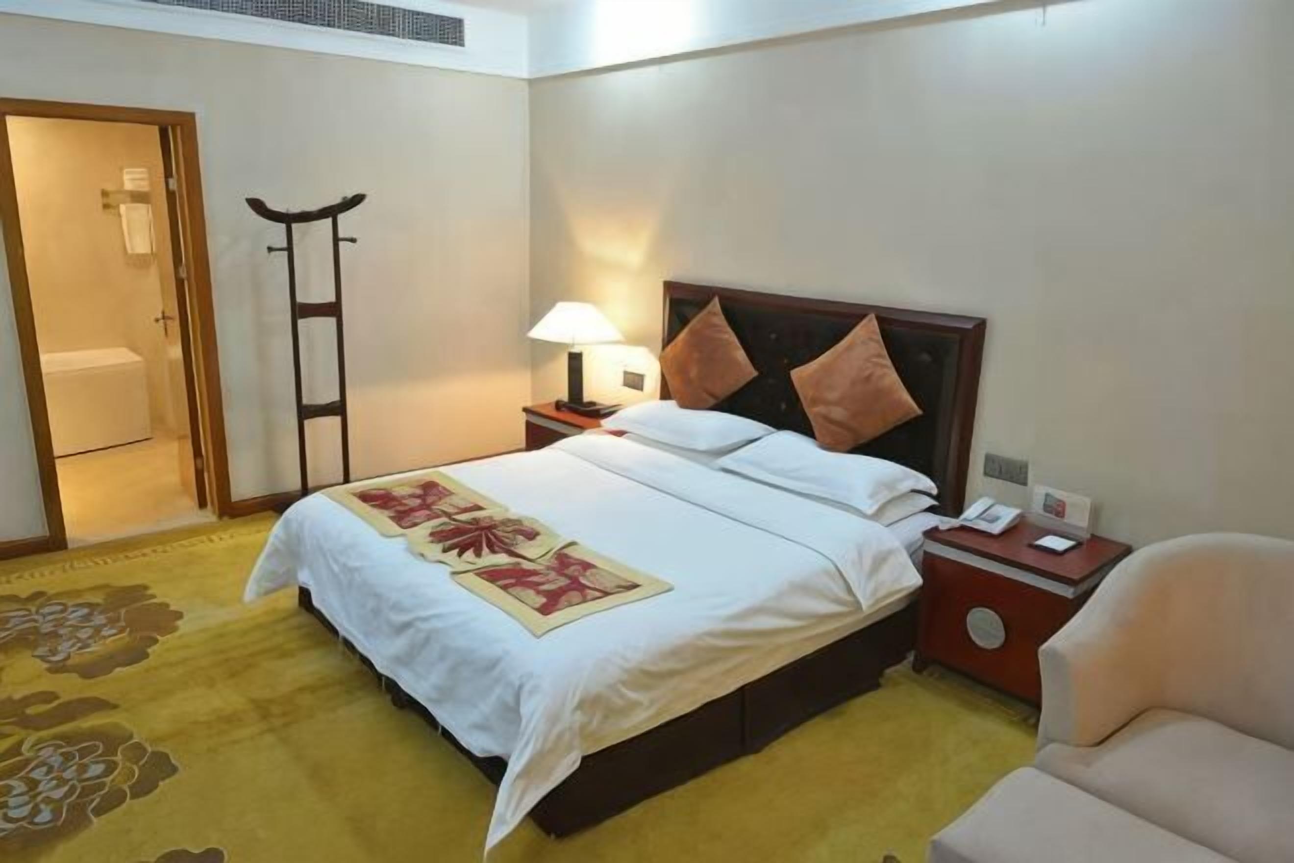 Una habitación de hotel con cama, mesita de noche, lámpara, silla y puerta que da a un baño.