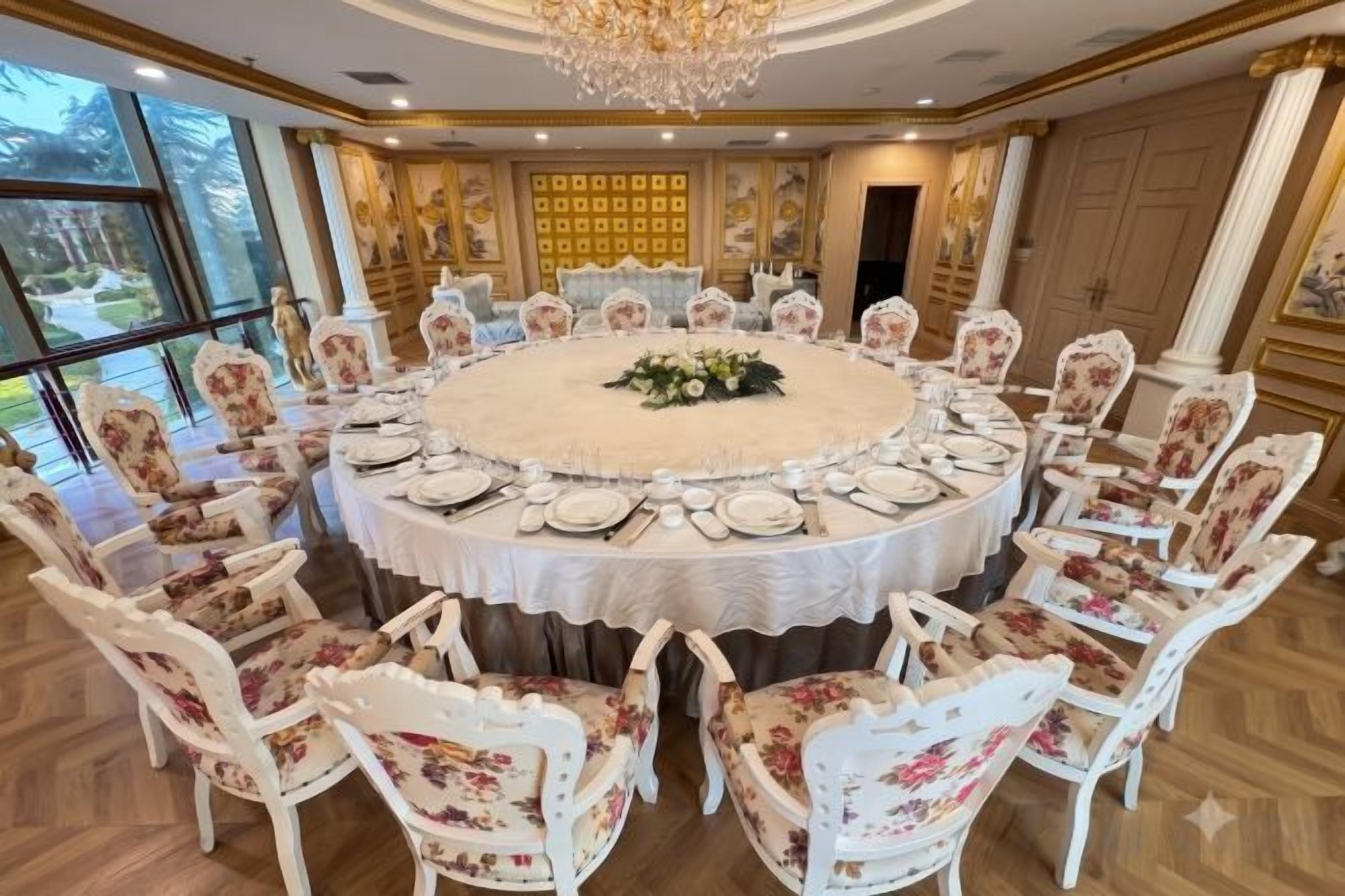 A round dining table with floral chairs, surrounded by a large window and a chandelier.