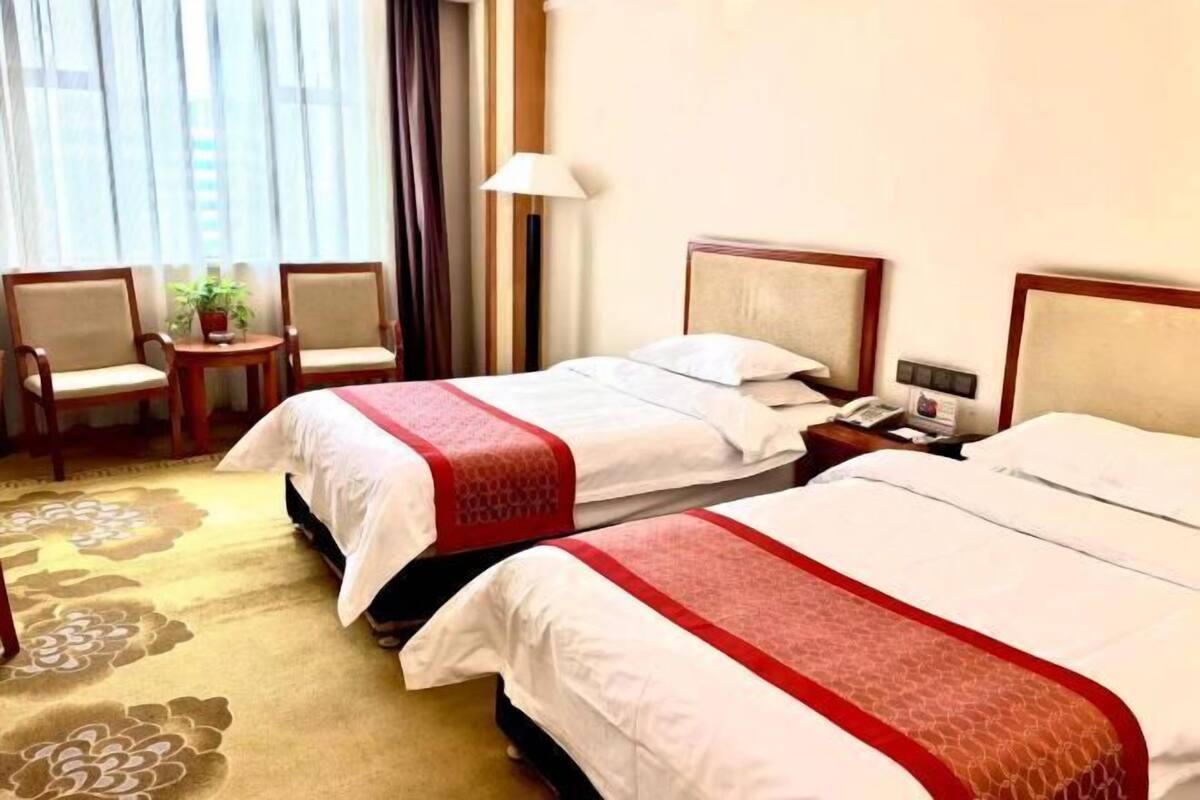 A hotel room with two beds, a small table with a plant, a lamp, and a view of the outside through large windows.
