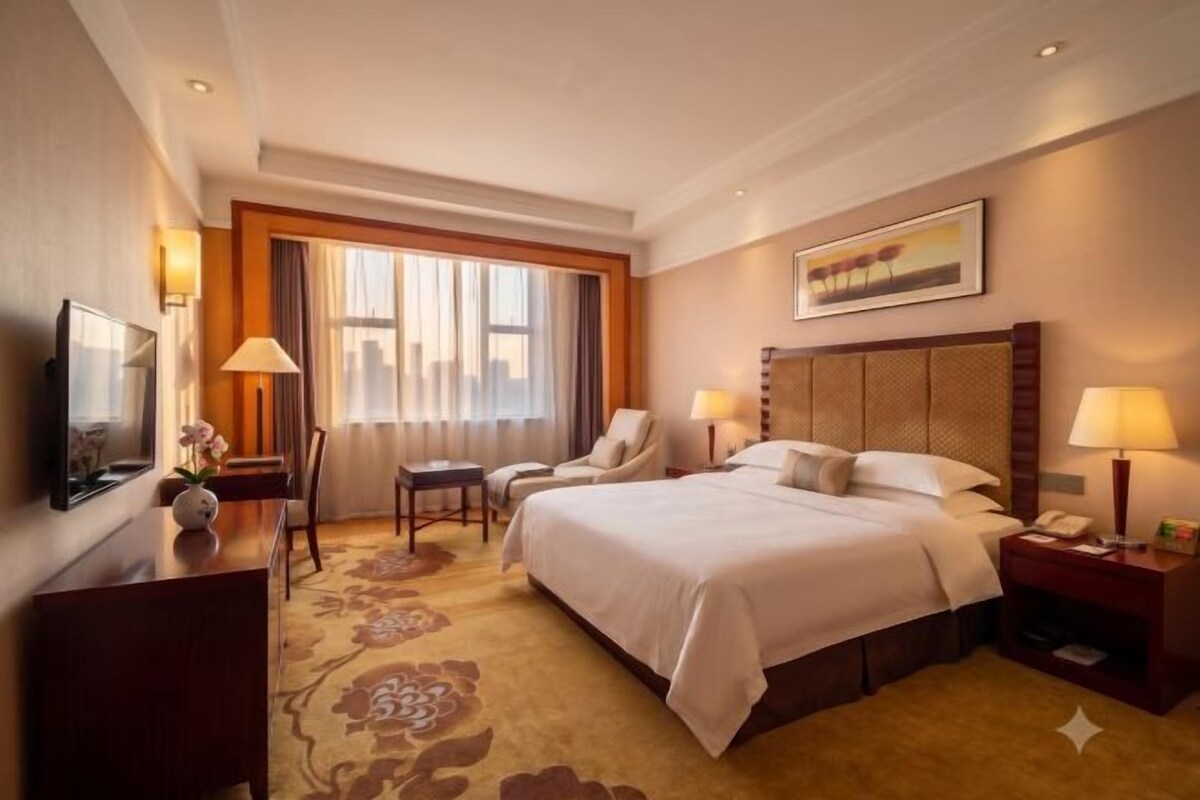 Une chambre d’hôtel avec un lit, un bureau, une chaise, une télévision et une fenêtre avec des rideaux.