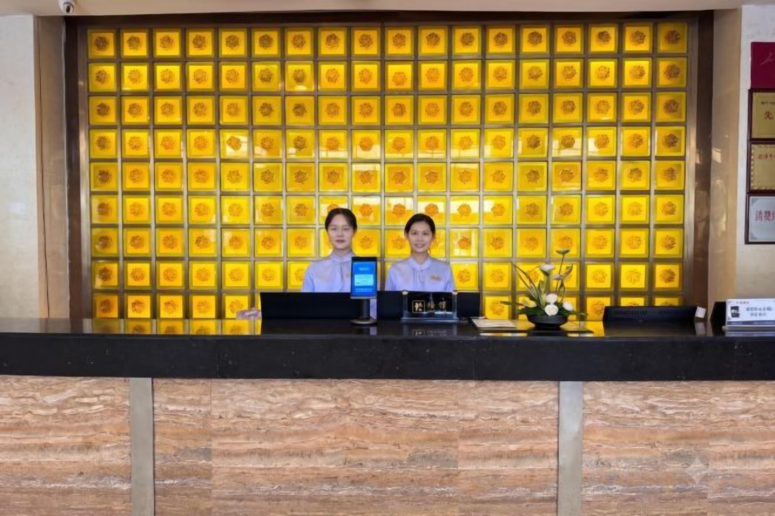 A reception desk with two staff members, a digital display, and a decorative wall with a pattern.