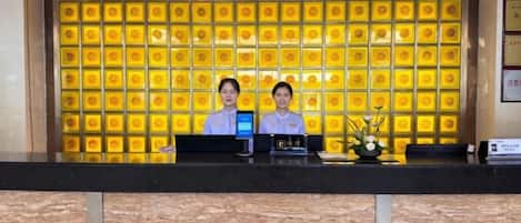 A reception desk with two staff members, a digital display, and a decorative wall with a pattern.