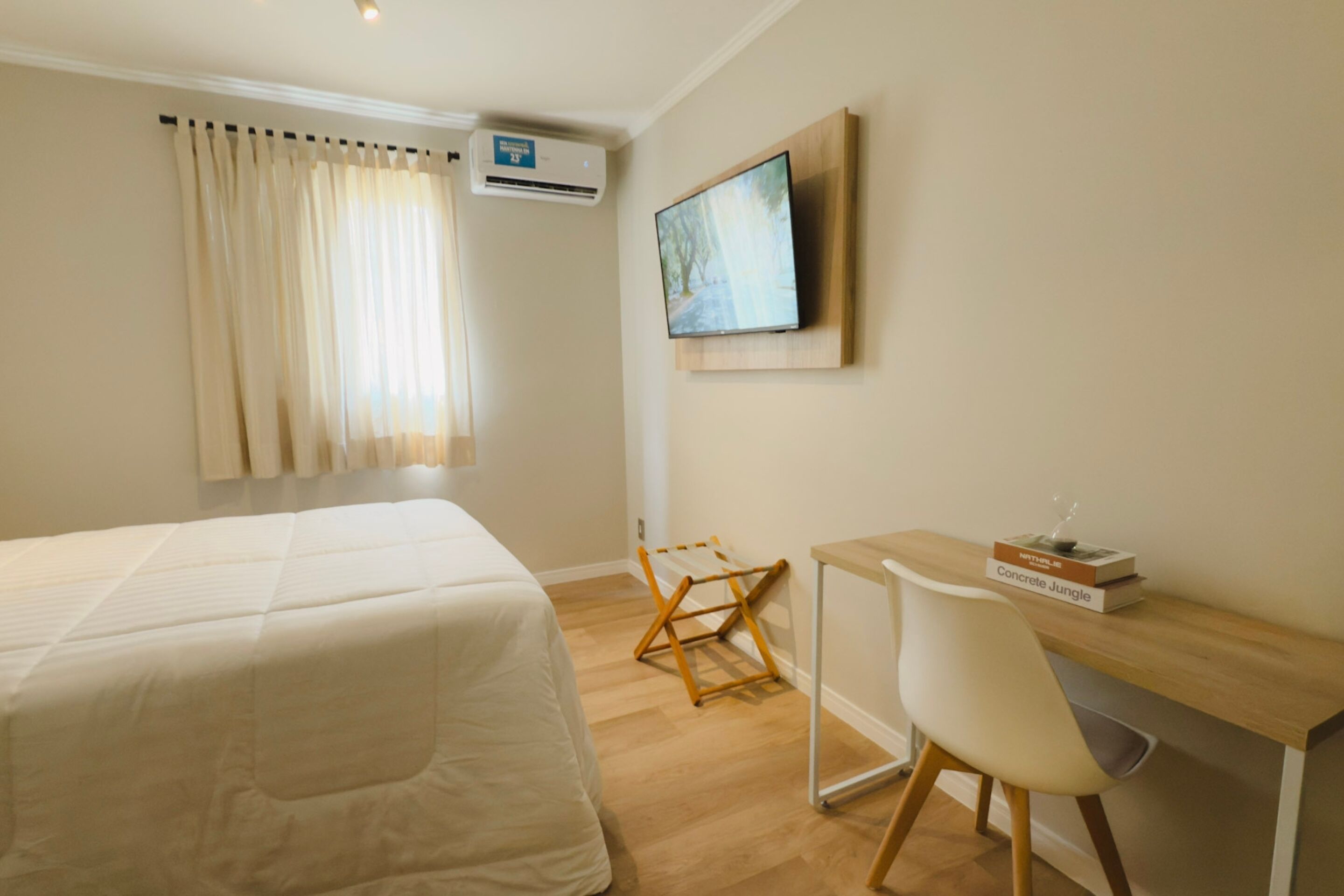 A hotel room with a bed, a desk with a chair, a television mounted on the wall, and a window with curtains.