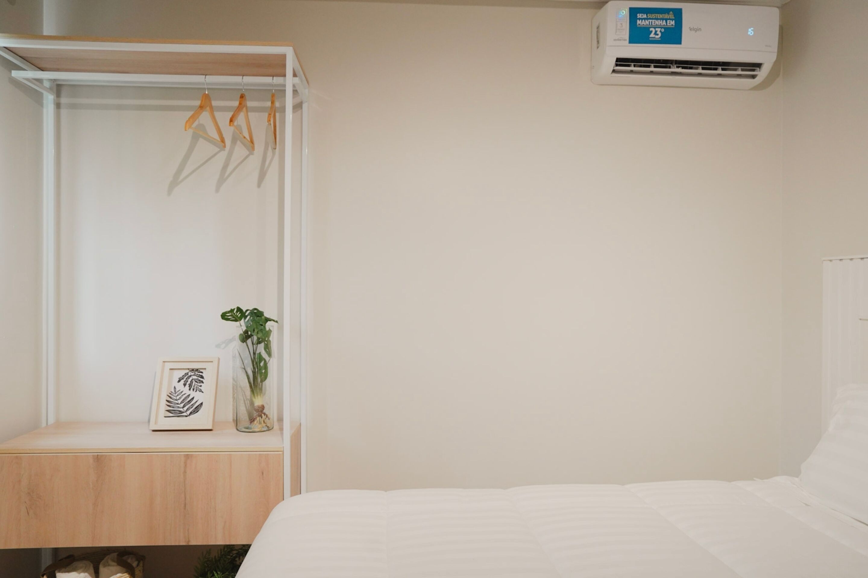 Ein Schlafzimmer mit Bett, Nachttisch, Kleiderstange, Klimaanlage und einem gerahmten Kunstwerk mit Blättern.