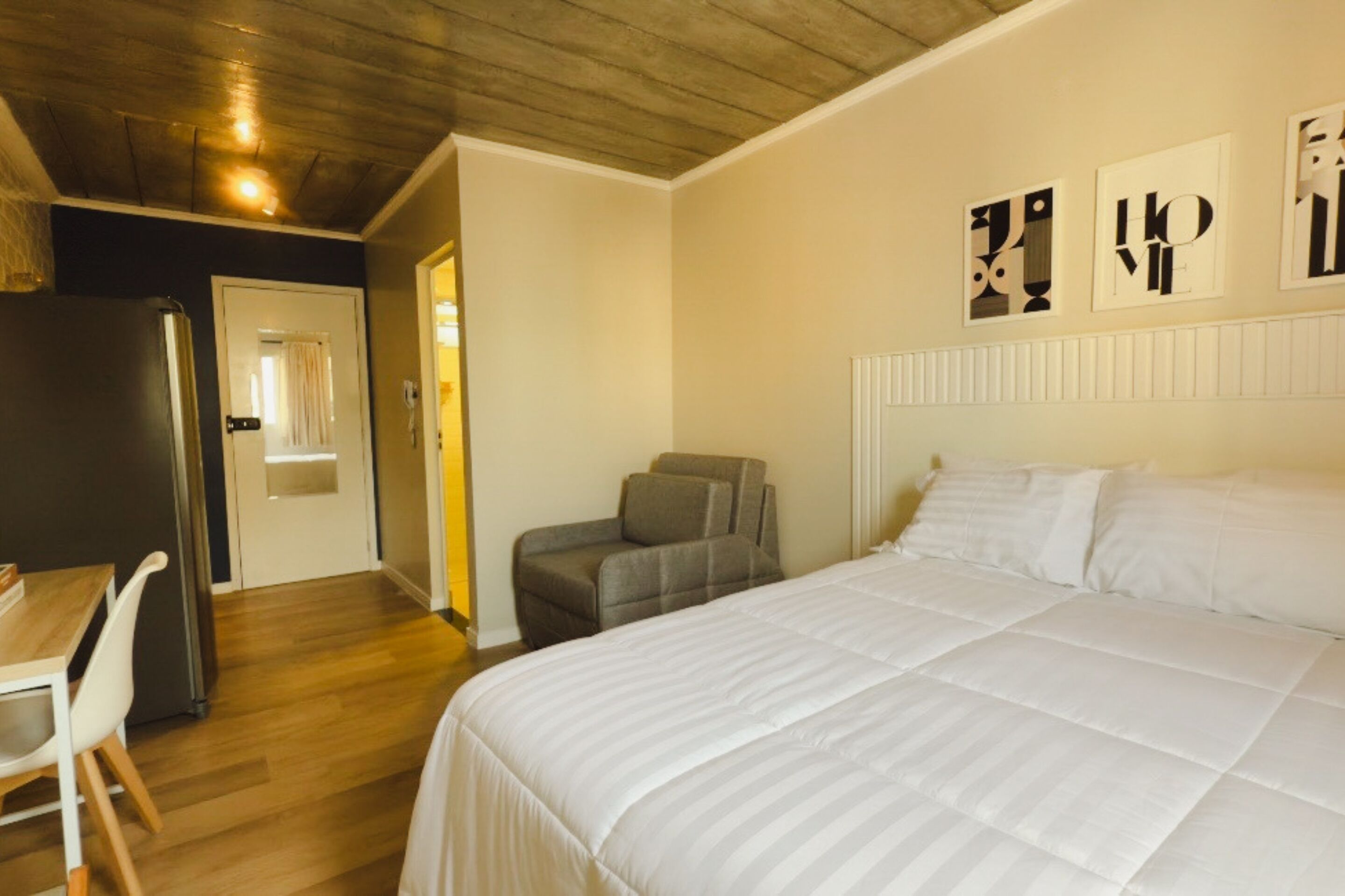 Een slaapkamer met een groot bed, een bureau en een stoel. Er is een koelkast en een deur naar een andere kamer.